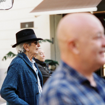 Man wearing a black hat and blue jacket walking outdoors with blurred people and an umbrella in the background.