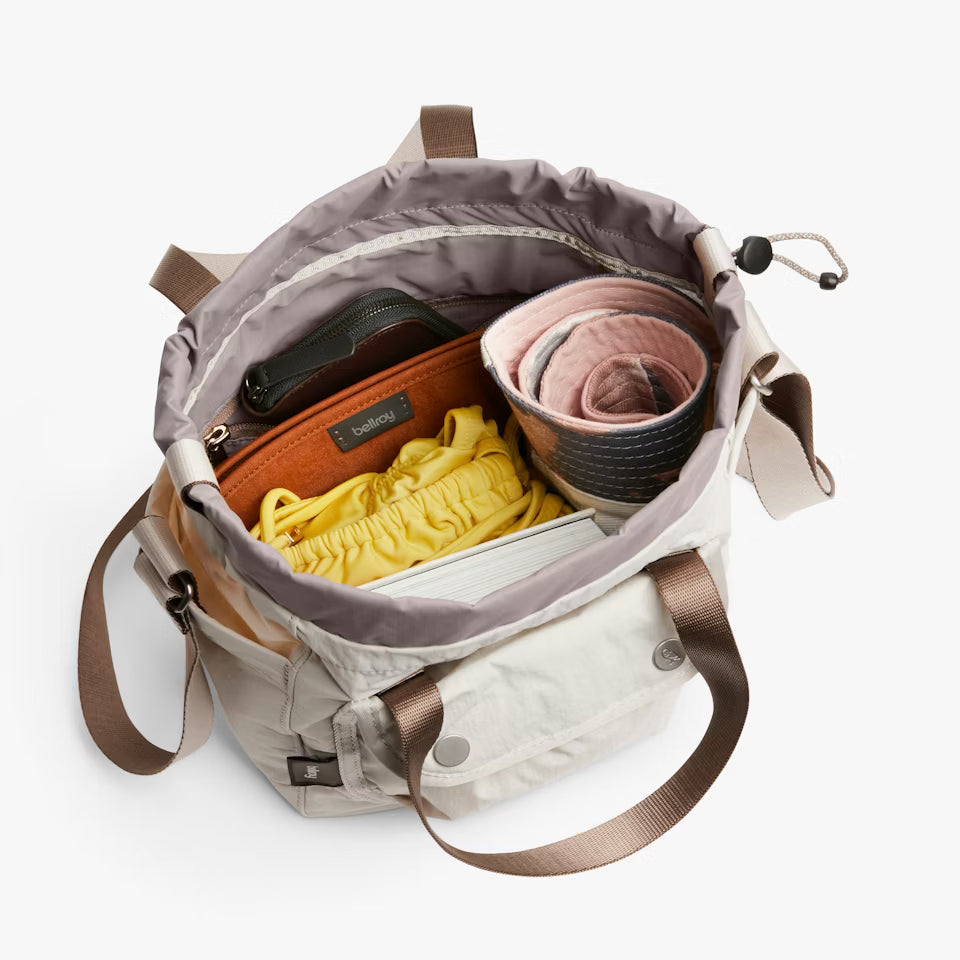 Open beige backpack with brown straps containing a suitcase, yellow garment, and pink item on a white background