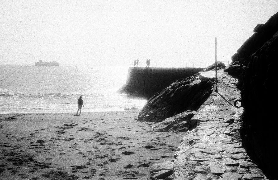 Grainy black and white image of a person on a rocky beach with a boat in the water and silhouetted people on a peer