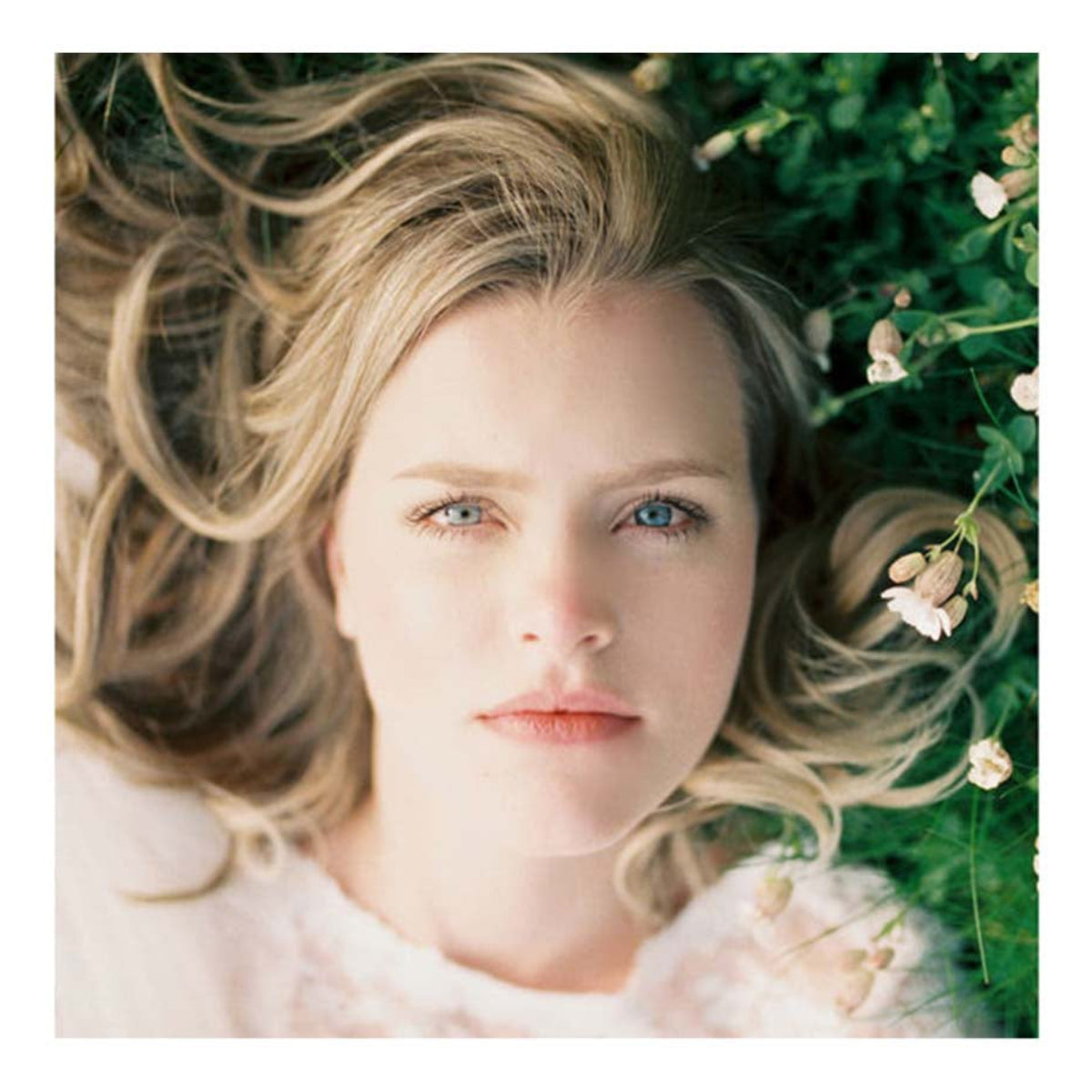 A square art print featuring a close-up portrait of a young blonde woman lying in green grass among small white flowers, centered on a white background.