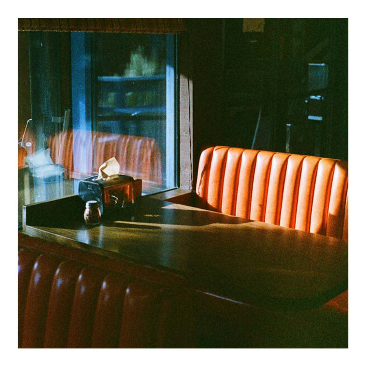 A square art print depicting a moody, warmly lit retro diner booth with a ribbed orange-brown leather seat and a dark wooden counter under dramatic window light.
