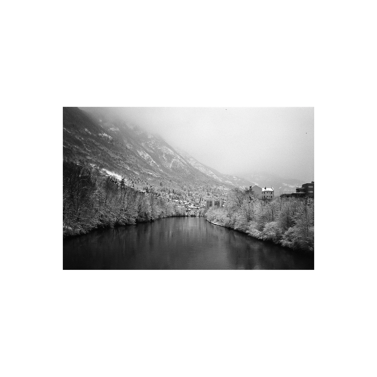 Black and white landscape photograph of a snowy mountain valley with a river in the foreground, snow-covered trees, a small town, and a building on the right.