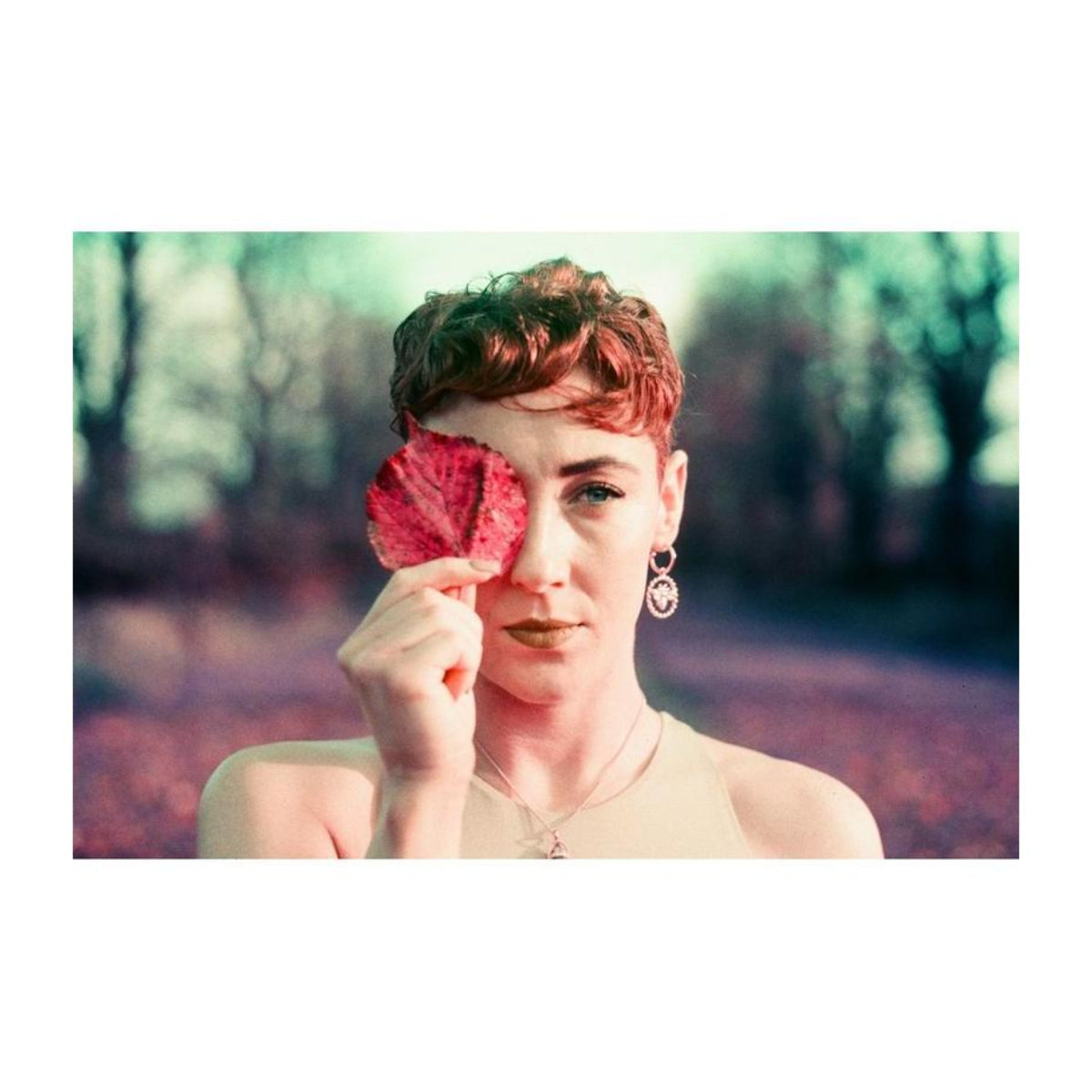 Person holding a red leaf over one eye, captured with vintage film tones and purple color shifts.