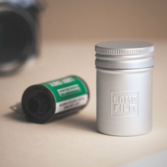 Roll of film with a green label next to tin container that says LOMO Film with a blurred camera in the background.