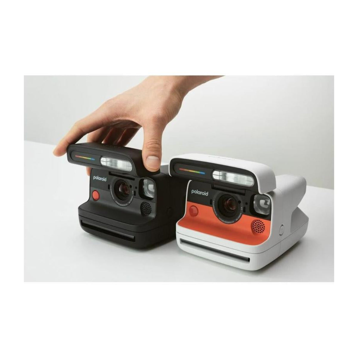 Hand adjusting a black Polaroid Flip instant camera next to a white and orange model on a white surface.