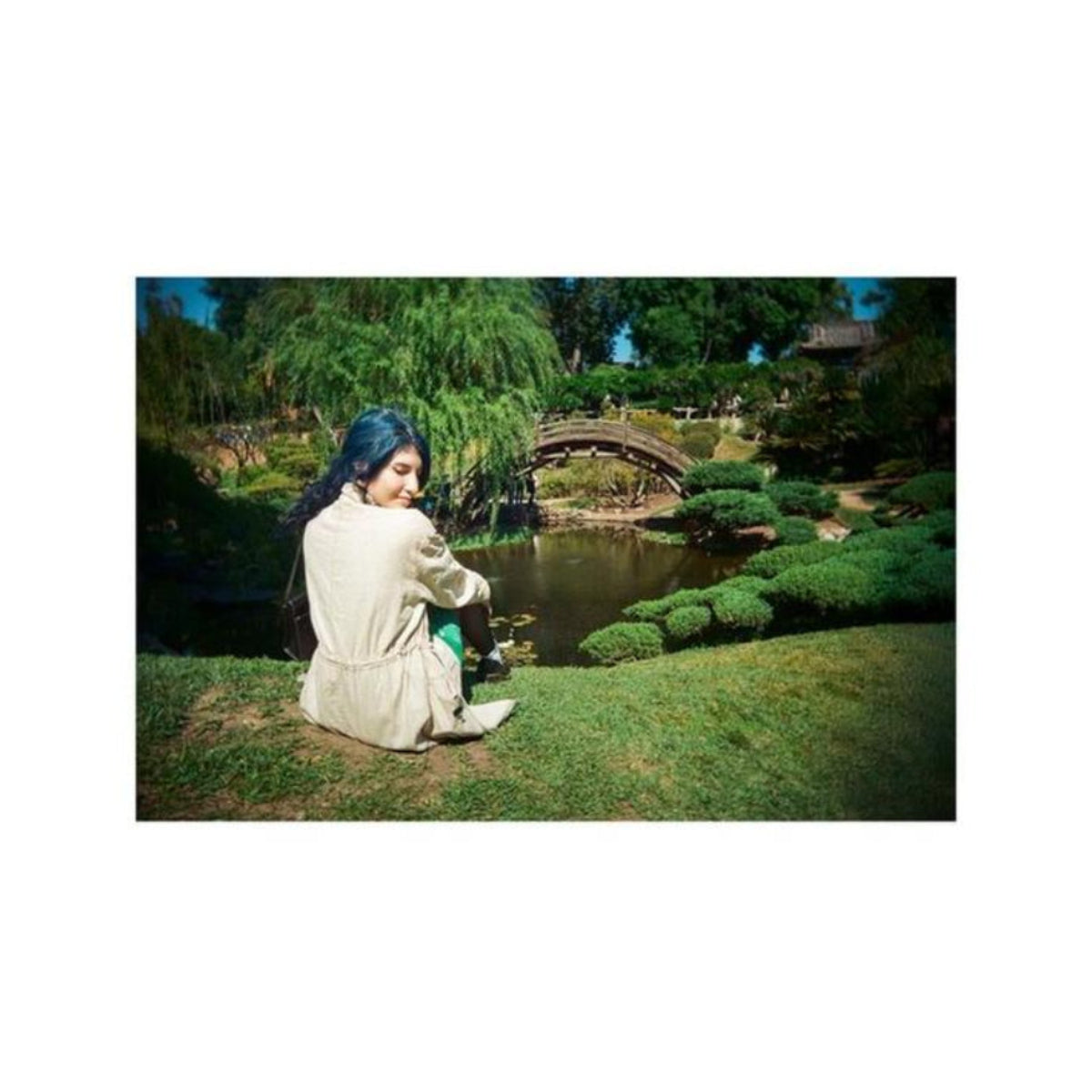 Panoramic shot of a woman in a beige coat sitting by a pond in a Japanese garden, captured with a 35mm film camera.