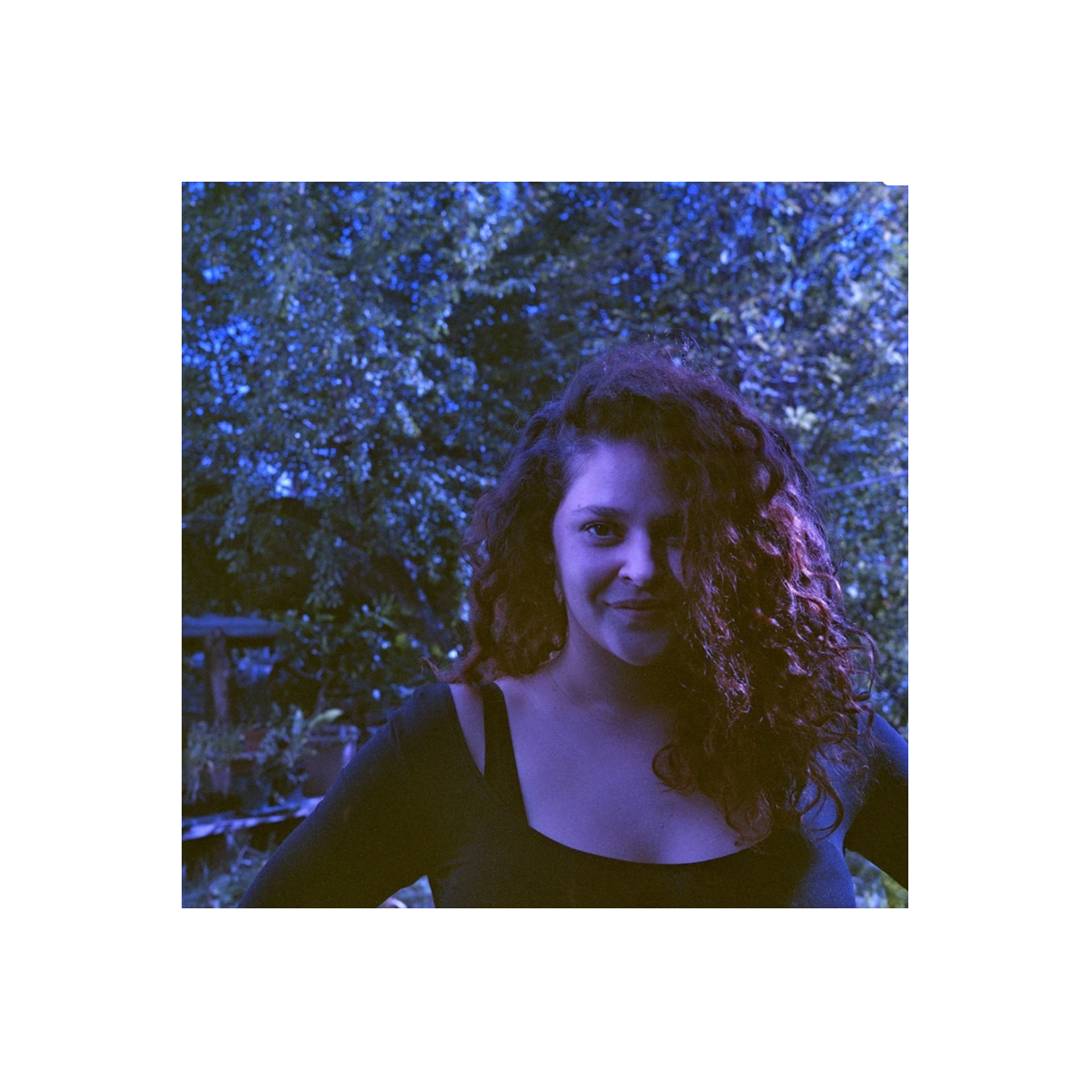 A woman with long, curly dark hair wearing a black top, photographed outdoors at night with blue-toned lighting against a background of trees.