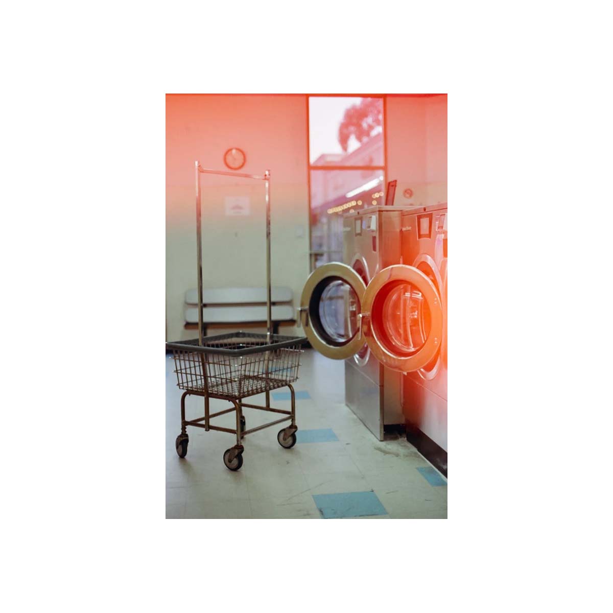A vintage metal laundry cart stands beside a stainless steel front-loading washing machine in a laundrette, with warm orange-red light reflecting off the machine and a faded red-orange gradient overlay across the photo.