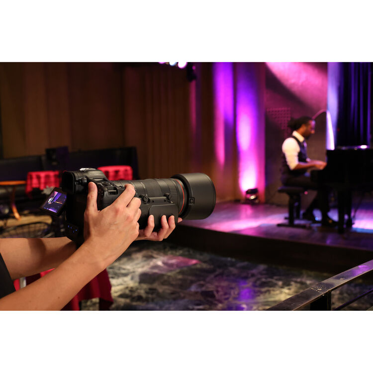 A close-up of a person holding a large black video camera with a long telephoto lens, with a blurred stage scene in the background featuring a pianist under purple stage lights.