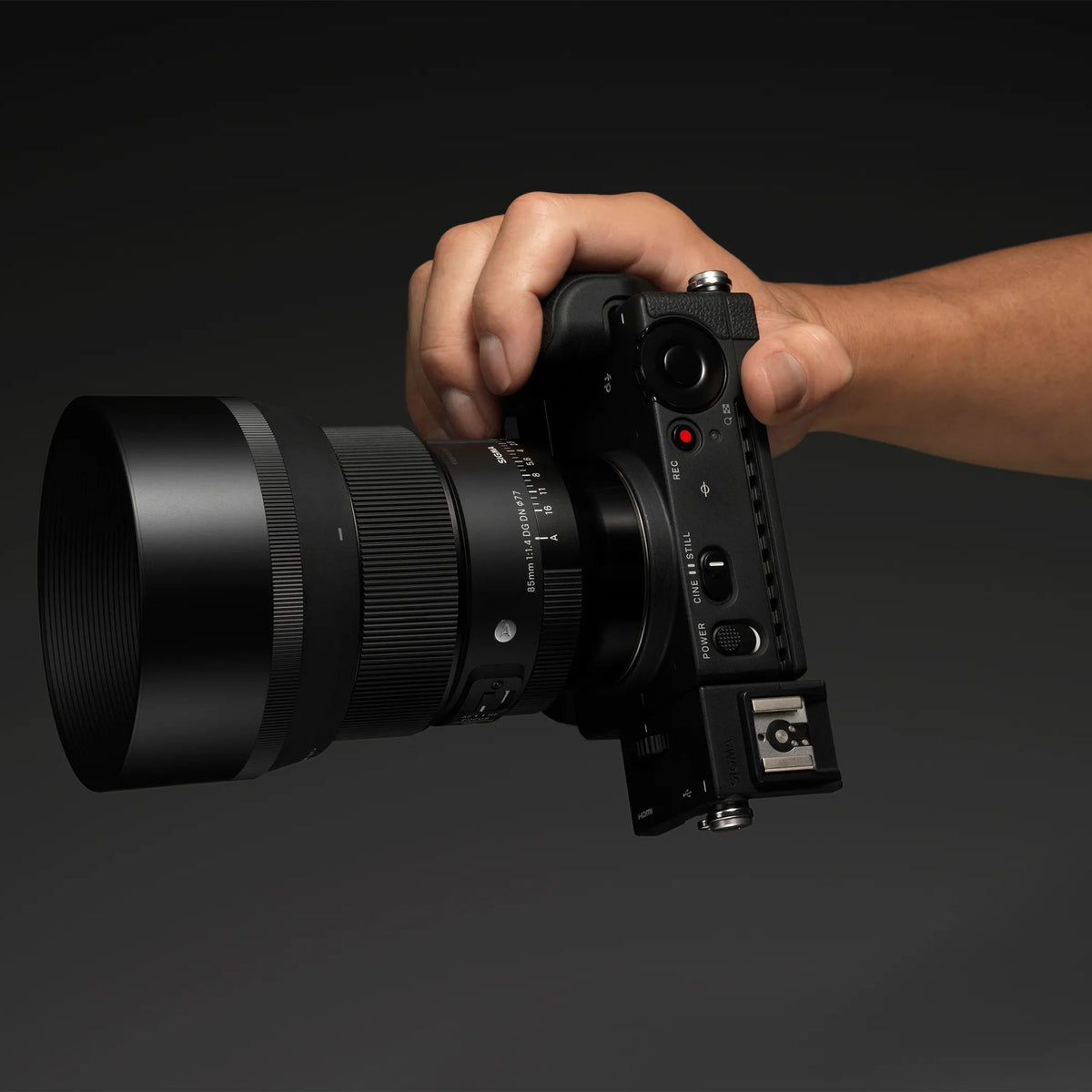 A hand holding a black film camera with a large lens attached, shown against a dark gray studio background.