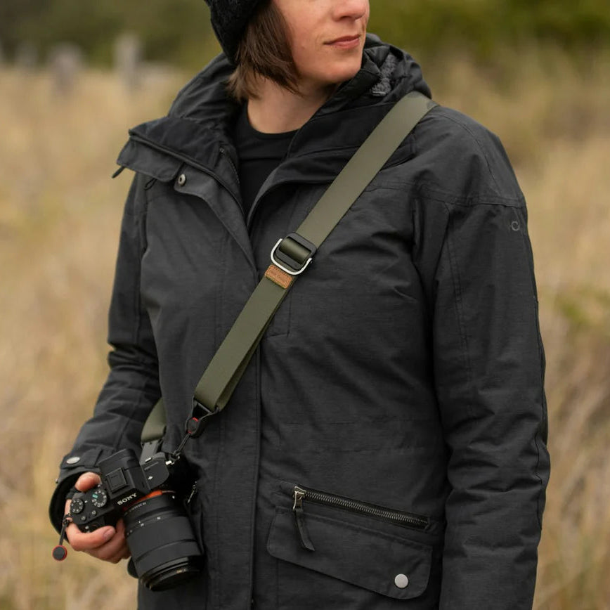 Person wearing a dark jacket with a camera on a strap, standing in a natural setting.