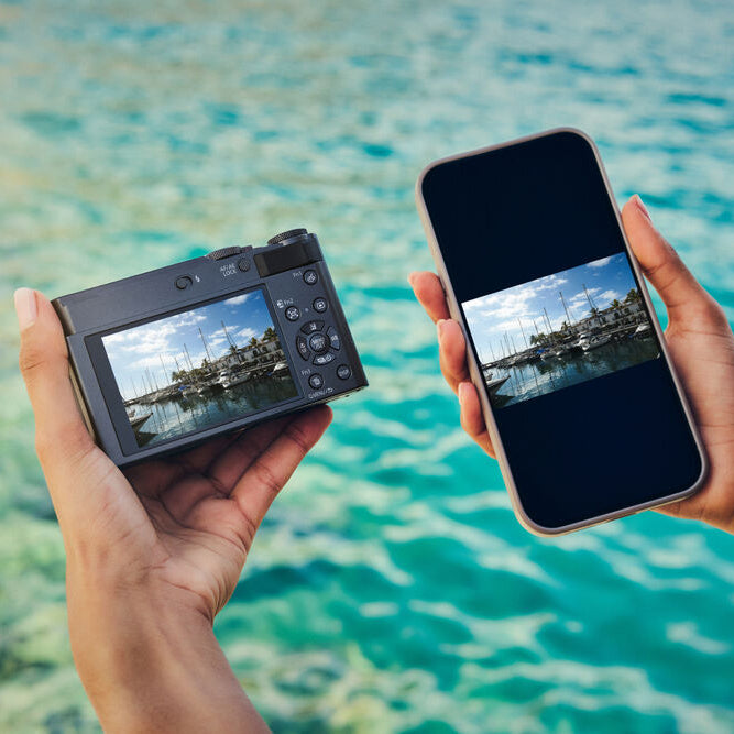 Two hands holding a camera and a smartphone displaying a scenic view of water and sky.