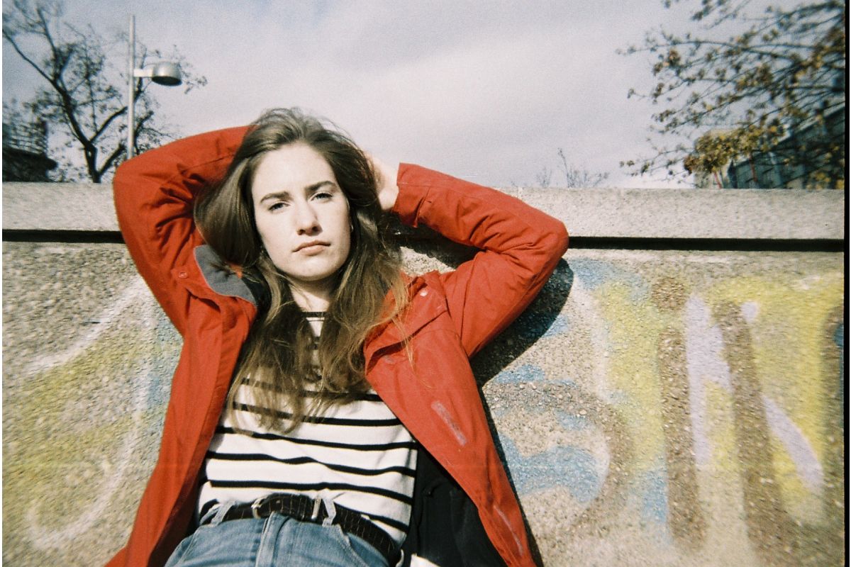 Person wearing a red jacket and striped shirt leaning against a wall with graffiti.