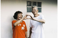 Two elderly people making a heart shape with their hands against a plain wall.