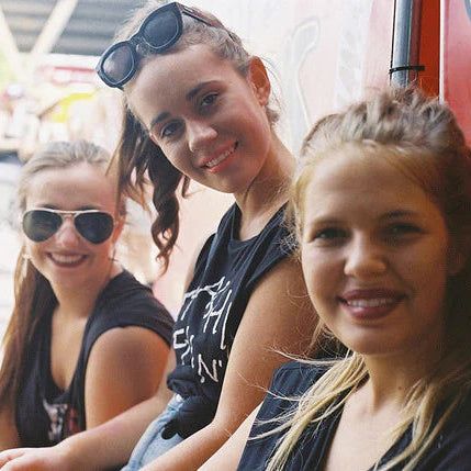 Three young women smiling and posing together outdoors, with one wearing black sunglasses on her head, another wearing black sunglasses on her face, and a third wearing a black graphic t-shirt. A bright red vehicle is visible in the background.