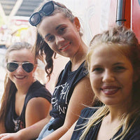 Three young women smiling and posing together outdoors, with one wearing black sunglasses on her head, another wearing black sunglasses on her face, and a third wearing a black graphic t-shirt. A bright red vehicle is visible in the background.