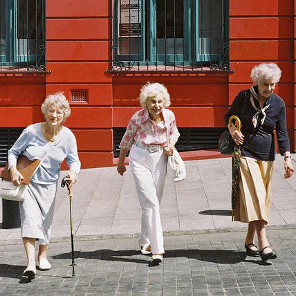 Three elderly women walking on a paved sidewalk in front of a bright red building facade. The woman on the left uses a cane and carries a brown handbag, the middle woman wears a pink and white patterned top with white trousers, and the woman on the right wears a navy cardigan, beige skirt, and black shoes.