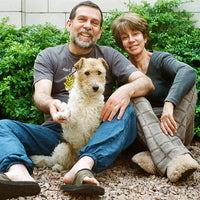 A smiling man and woman sitting outdoors on a gravel surface in front of green bushes, with a small white and tan dog sitting between them. The man wears a dark gray graphic T-shirt, blue jeans, and sandals, while the woman wears a dark long-sleeve top and gray slip-on shoes.