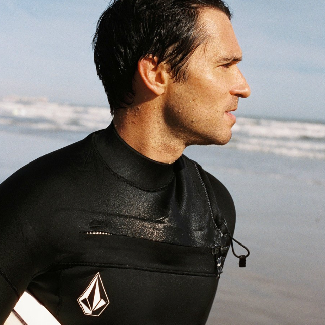 Man in a black wetsuit holding a surfboard on a beach