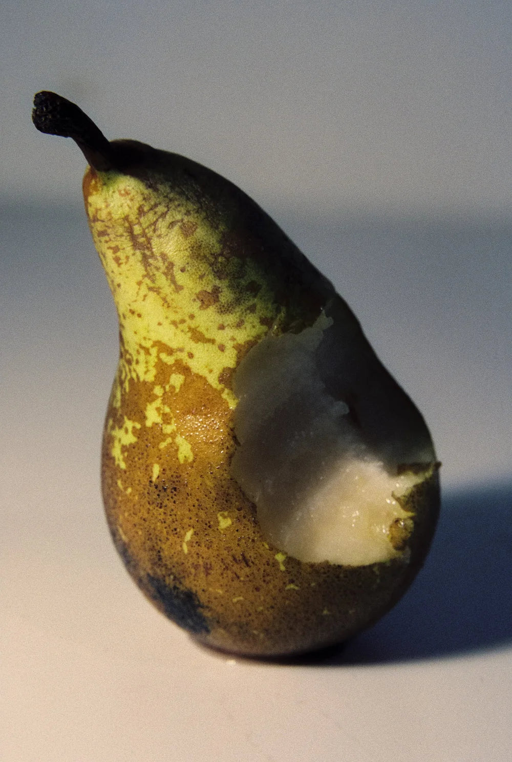 A single pear photographed on a plain light background, with one side bitten to show the white flesh. The rind is mottled yellow-green and brown.