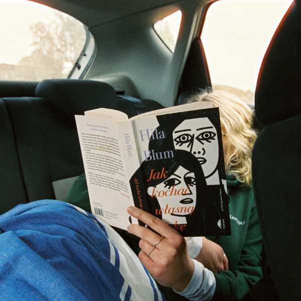 A person sitting in a car backseat reads a book titled 'Jak kochaŃ wasłszaŃ' by Hila Blum, with the book cover featuring a black-and-white illustrated face and orange text.