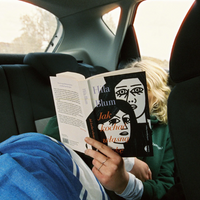 A person sitting in a car backseat reads a book titled 'Jak kochaŃ wasłszaŃ' by Hila Blum, with the book cover featuring a black-and-white illustrated face and orange text.