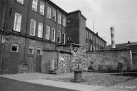 Black and white photograph of an industrial brick courtyard with a graffiti-covered wall and a round concrete structure in the foreground, with tall brick buildings and a striped chimney in the background.