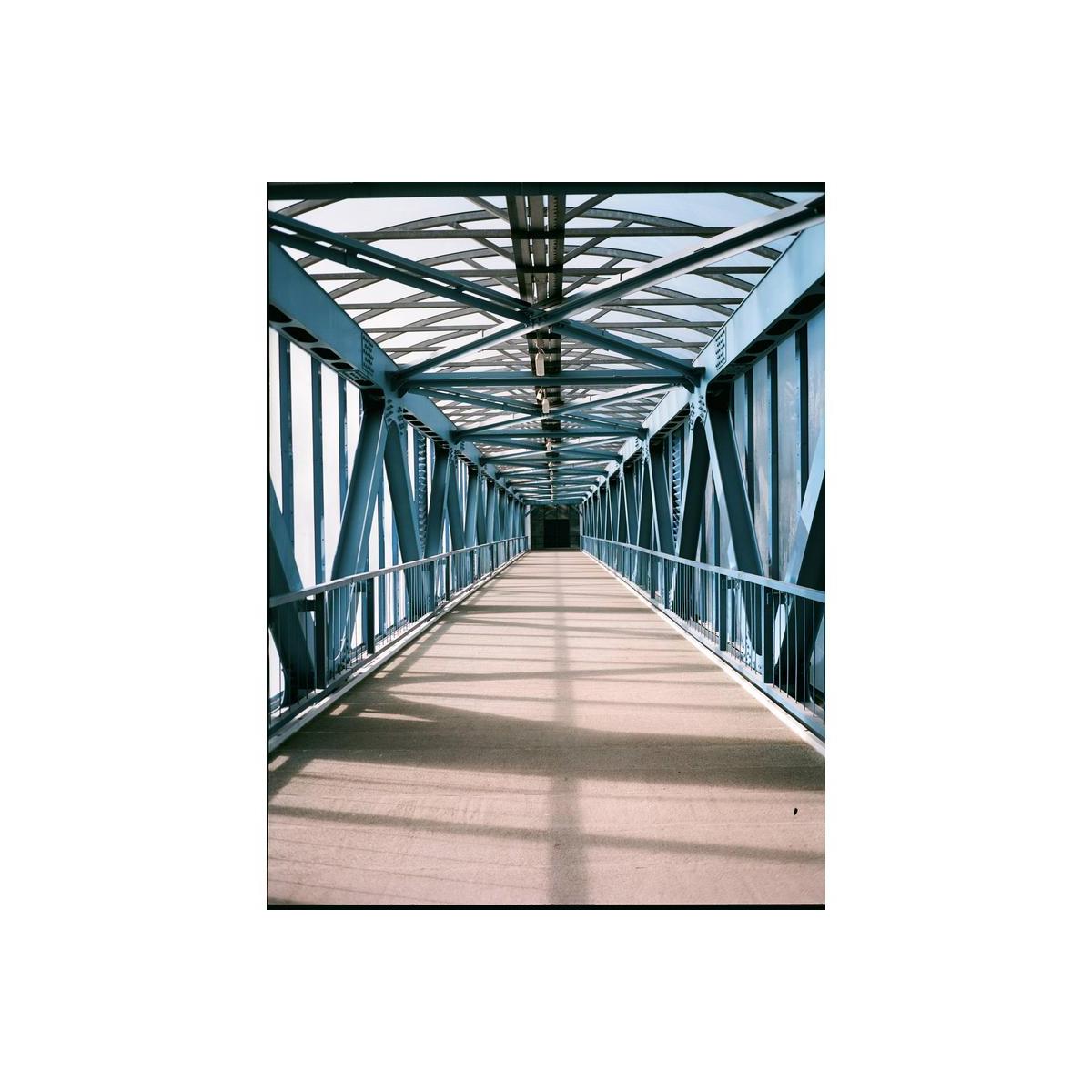 A perspective view down a covered bridge walkway with a blue steel truss structure and a beige floor, framed by repeating blue beams.