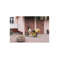 A yellow electric scooter with a delivery box on the back is parked on a sidewalk in front of a building with brown doors and a planter box in the foreground.
