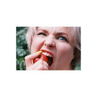A close-up lifestyle shot of a woman with short blonde hair biting into a red apple, holding a yellow object near her mouth, with a blurred green outdoor background.