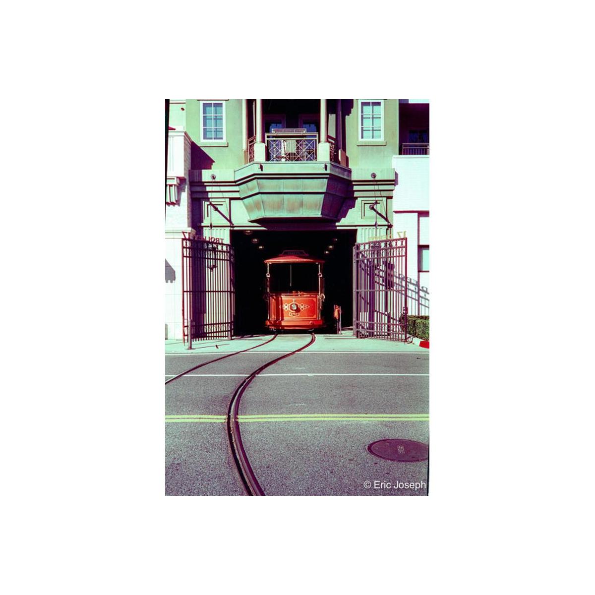 A vintage red cable car emerges from a dark tunnel beneath a green building, with curved tracks in the foreground and open metal gates on both sides.