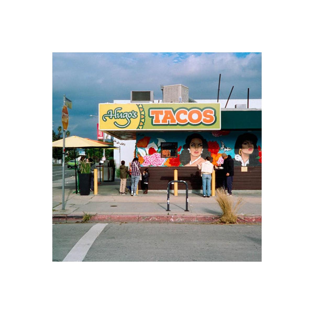 A street-side view of Hinga's Tacos with a colorful mural on the building, people standing in front, umbrellas on the sidewalk, and a cloudy sky overhead.