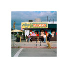 A street-side view of Hinga's Tacos with a colorful mural on the building, people standing in front, umbrellas on the sidewalk, and a cloudy sky overhead.