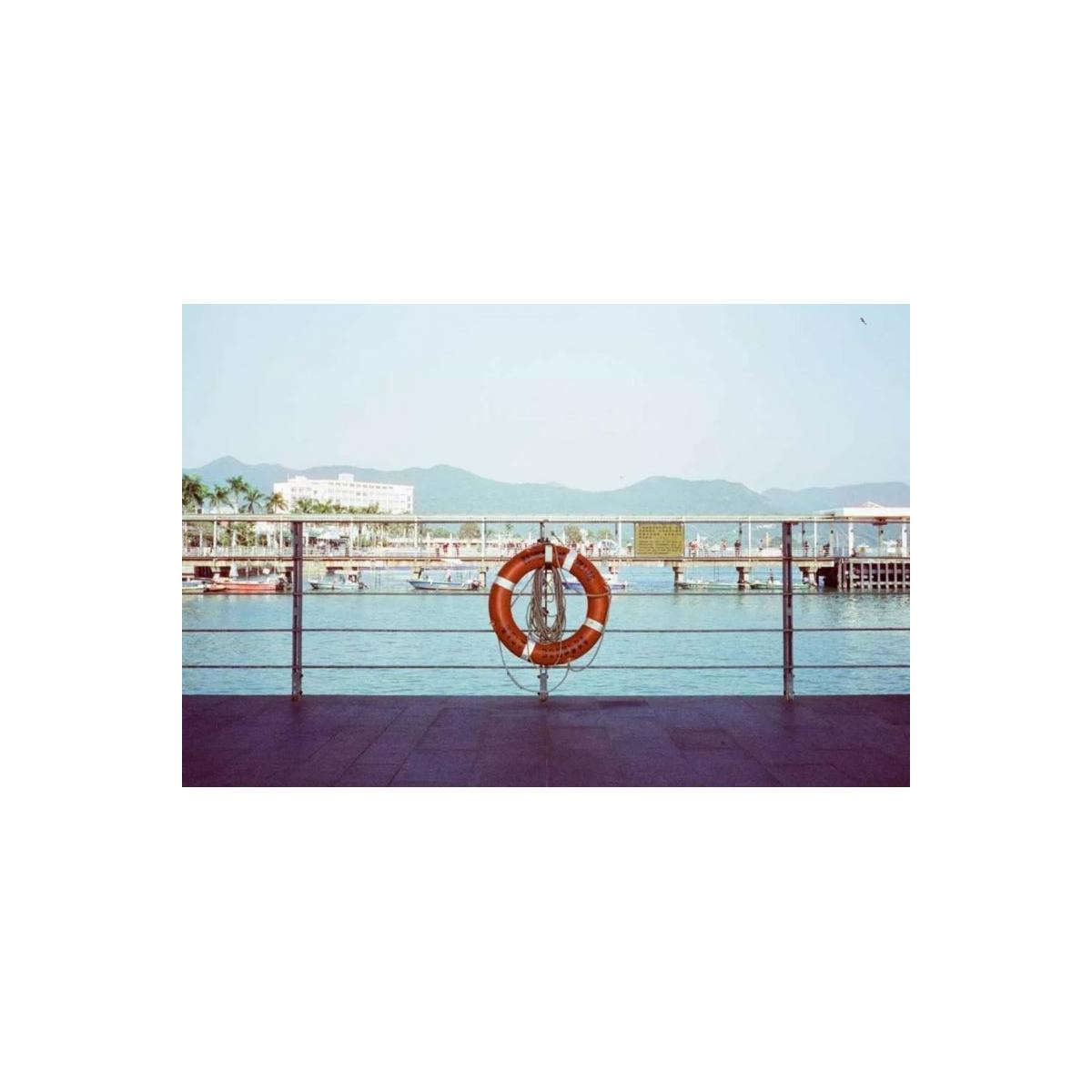 An orange life preserver with white stripes hangs on a metal railing overlooking a harbor with boats, water, and distant mountains under a pale blue sky.