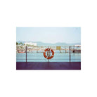An orange life preserver with white stripes hangs on a metal railing overlooking a harbor with boats, water, and distant mountains under a pale blue sky.