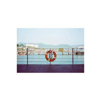 An orange life preserver with white stripes hangs on a metal railing overlooking a harbor with boats, water, and distant mountains under a pale blue sky.