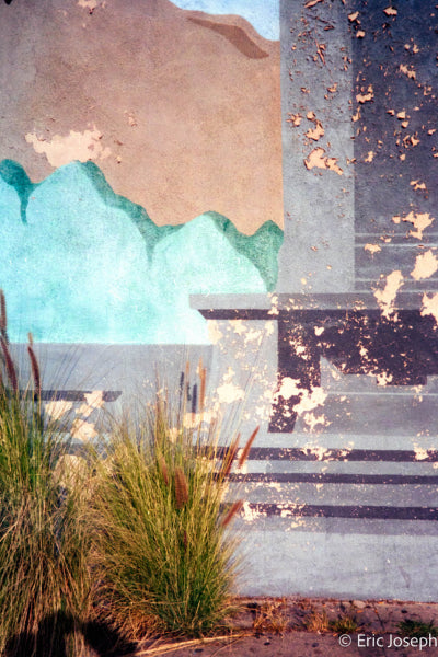 An abstract architectural photograph featuring a textured stucco wall with a painted turquoise wave design, a gray column, steps, and foreground grasses under warm sunlight. Signed by Eric Joseph in the bottom right corner.