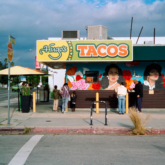 A street-side view of Hinga's Tacos with a colorful mural on the building, people standing in front, umbrellas on the sidewalk, and a cloudy sky overhead.