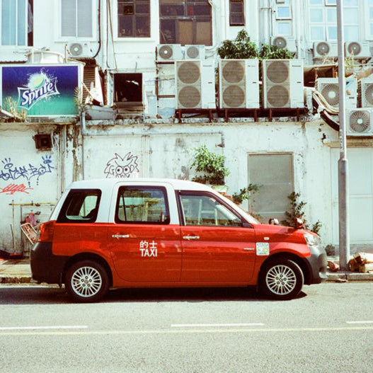 A red and white London-style taxi parked on a city street in front of a weathered white building with graffiti, air conditioning units, and a visible Sprite sign.