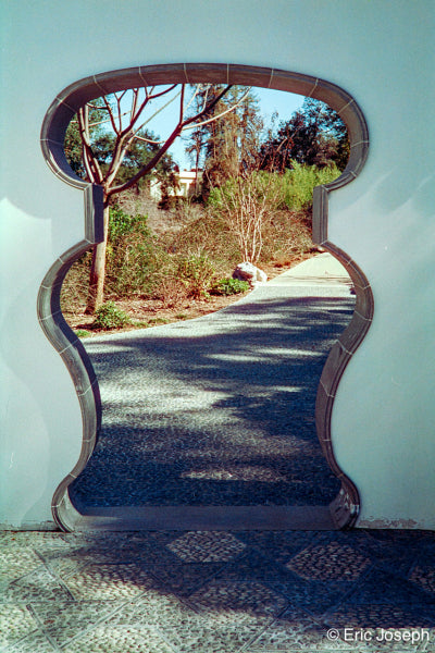 A decorative stone archway or doorway framing a sunlit gravel path with trees and shrubs beyond, set against a pale blue wall.