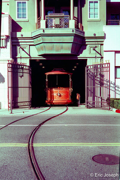 A vintage red cable car emerges from a dark tunnel beneath a green building, with curved tracks in the foreground and open metal gates on both sides.