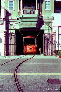 A vintage red cable car emerges from a dark tunnel beneath a green building, with curved tracks in the foreground and open metal gates on both sides.
