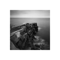 Black and white landscape photograph of a rocky coastal cliff overlooking a calm sea under an overcast sky.