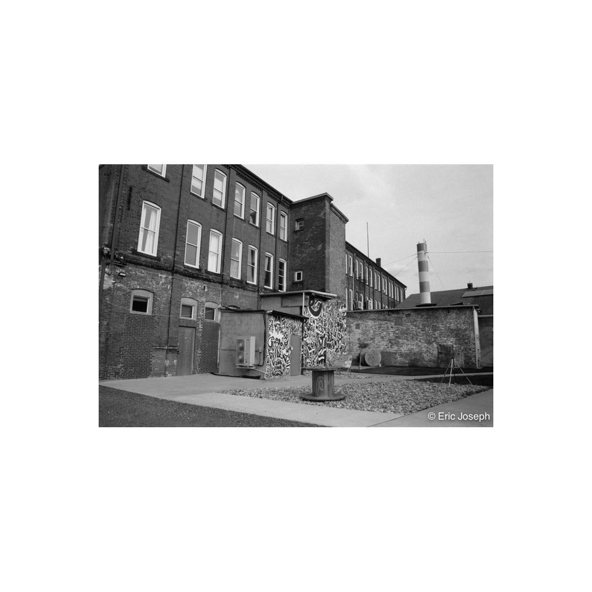 Black and white photograph of an industrial brick courtyard with a graffiti-covered wall and a round concrete structure in the foreground, with tall brick buildings and a striped chimney in the background.