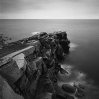Black and white landscape photograph of a rocky coastal cliff overlooking a calm sea under an overcast sky.