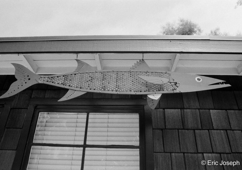 A large fish-shaped metal wall hanging with a spotted pattern, mounted under the eaves of a house above a window.