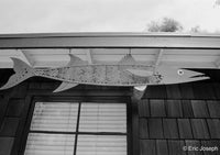 A large fish-shaped metal wall hanging with a spotted pattern, mounted under the eaves of a house above a window.
