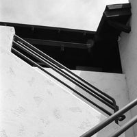 Black and white architectural photograph of an outdoor staircase with dark metal railings against a textured white wall and dark roofline.