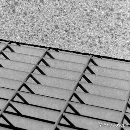 Black and white close-up photograph of textured concrete or stone tiles meeting a slatted metal grate, with a photographer's copyright watermark in the bottom right corner.