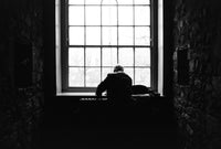 A black and white photograph of a person sitting in silhouette at a piano in a dimly lit stone room, facing a large multi-paned window.